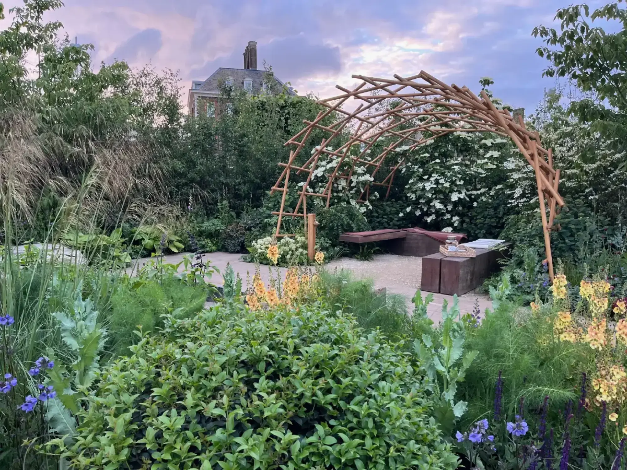 A small show garden with a curved woven timber archway over wooden benches and a gravel path, bordered by green plants and white, yellow, and blue flowers.