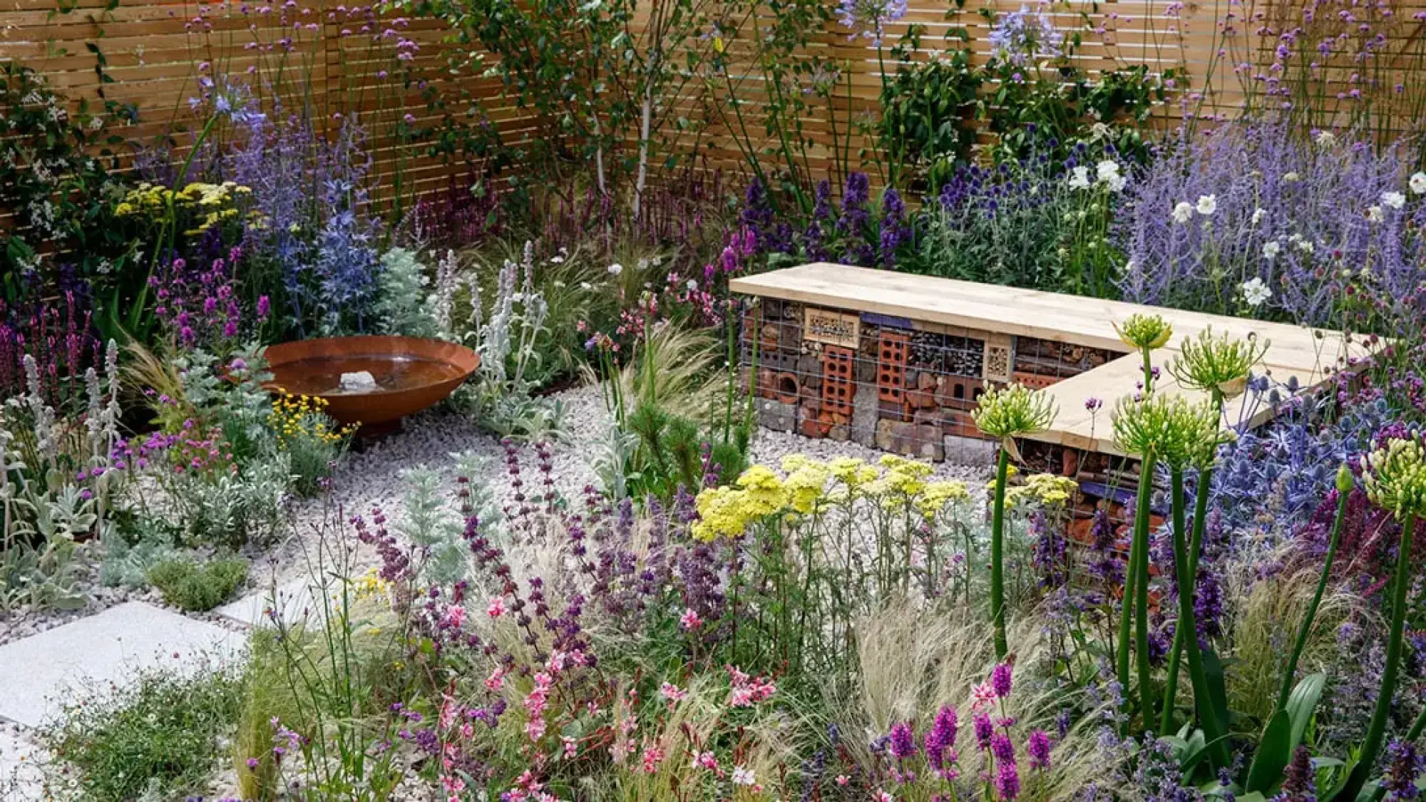 A vibrant, wildlife-friendly garden with a bug hotel bench, rusty bird bath, gravel paths, and mixed purple, pink, blue, yellow flowers.