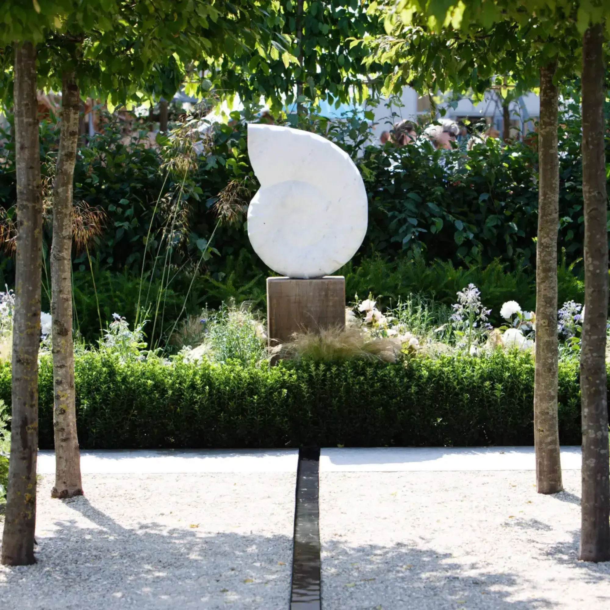 A contemporary garden featuring a white spiral sculpture, a dark central rill, and lush green planting with white and pale flowers, framed by mature trees.