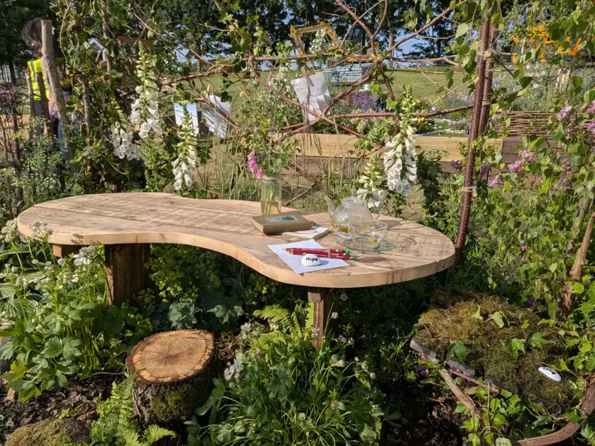 A rustic garden border features a curved timber table, a tree stump seat, white foxgloves, and a woven branch screen with pinned notes.