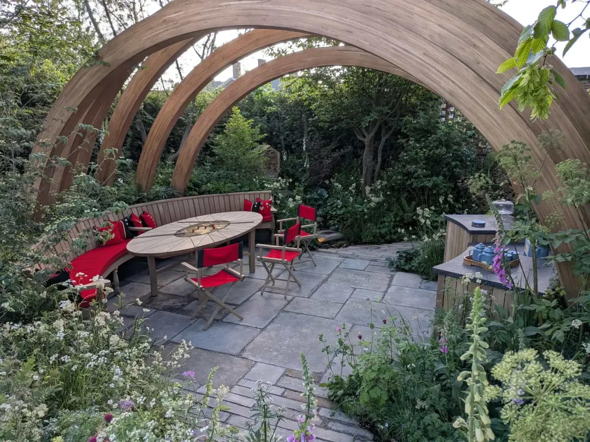 A small show garden featuring a striking arched timber structure, a curved wooden bench with red cushions, an oval dining table with a central fire pit, and stone paving, surrounded by lush, naturalistic planting with white and purple flowers.