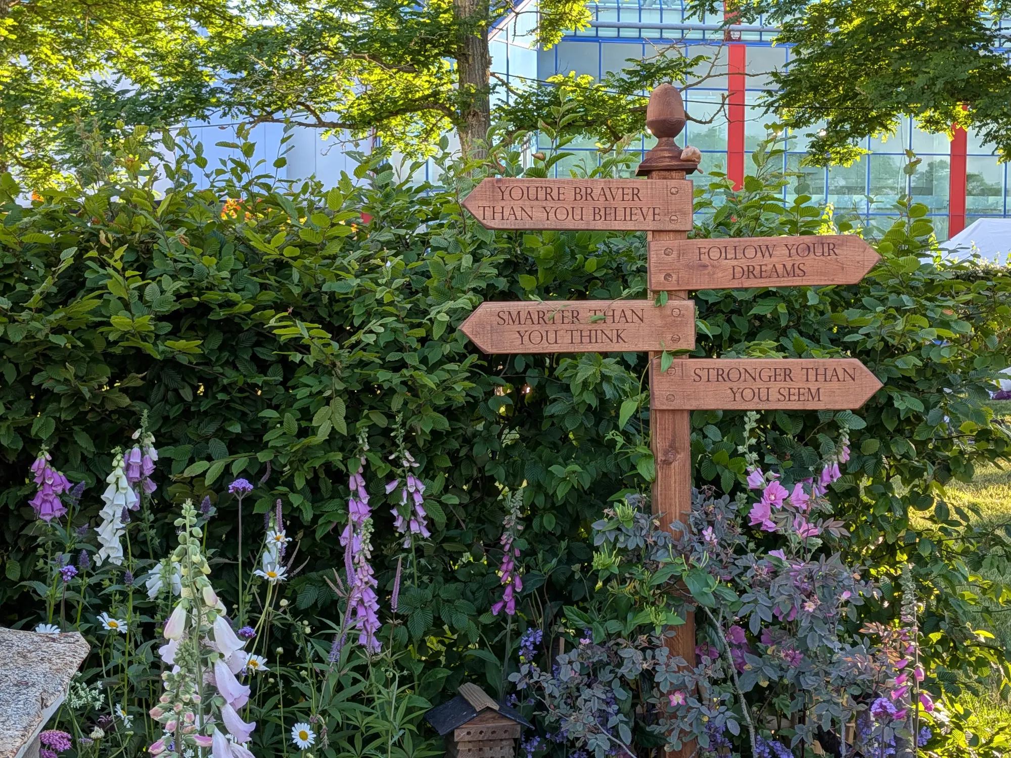A beautiful border features a wooden signpost with motivational quotes among a variety of foxgloves, daisies, and mixed green and purple foliage, with a small insect hotel at the base.
