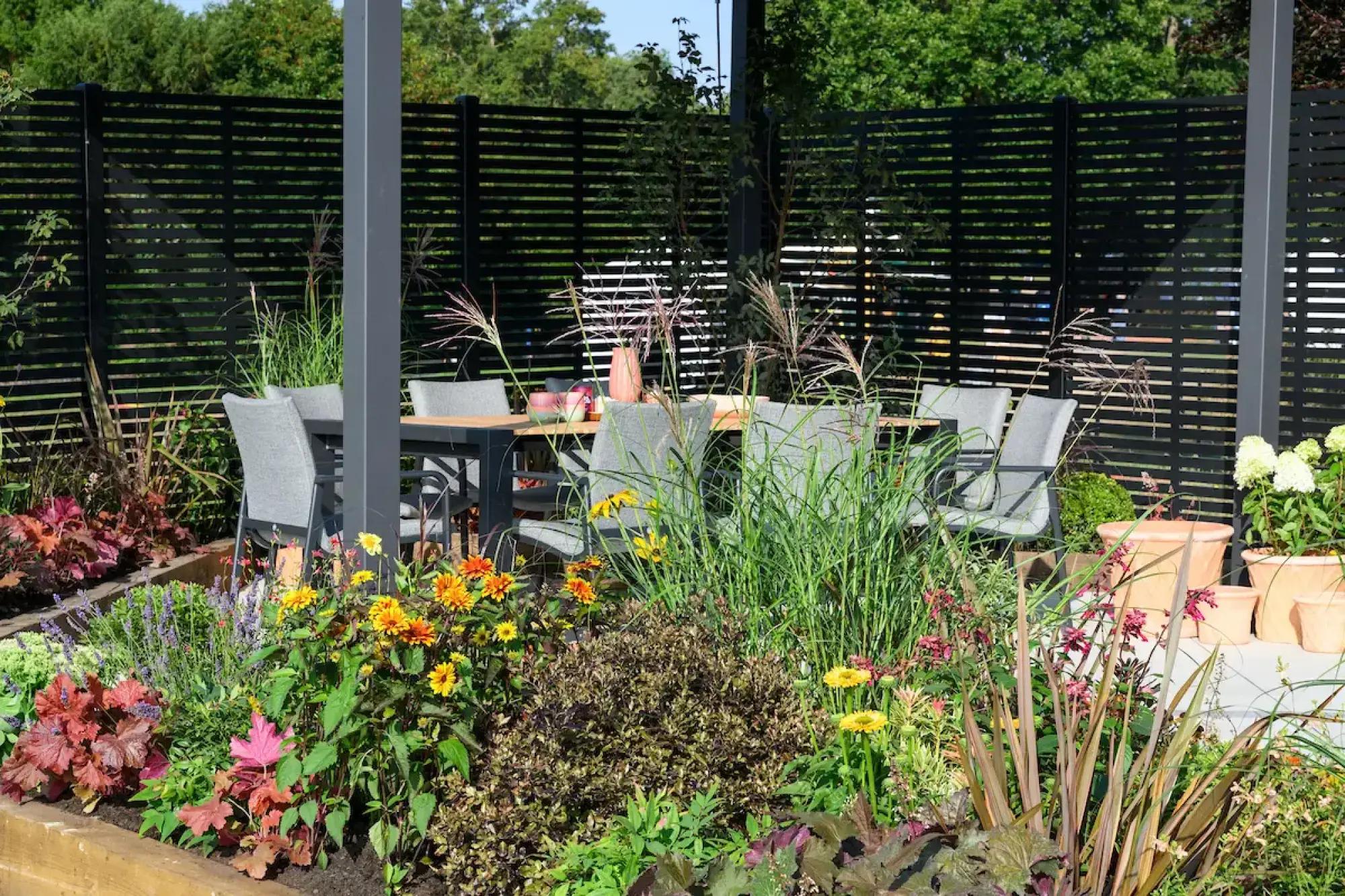 A contemporary garden showcases a dark grey pergola, slatted fence, outdoor dining, and raised beds bursting with colourful planting.