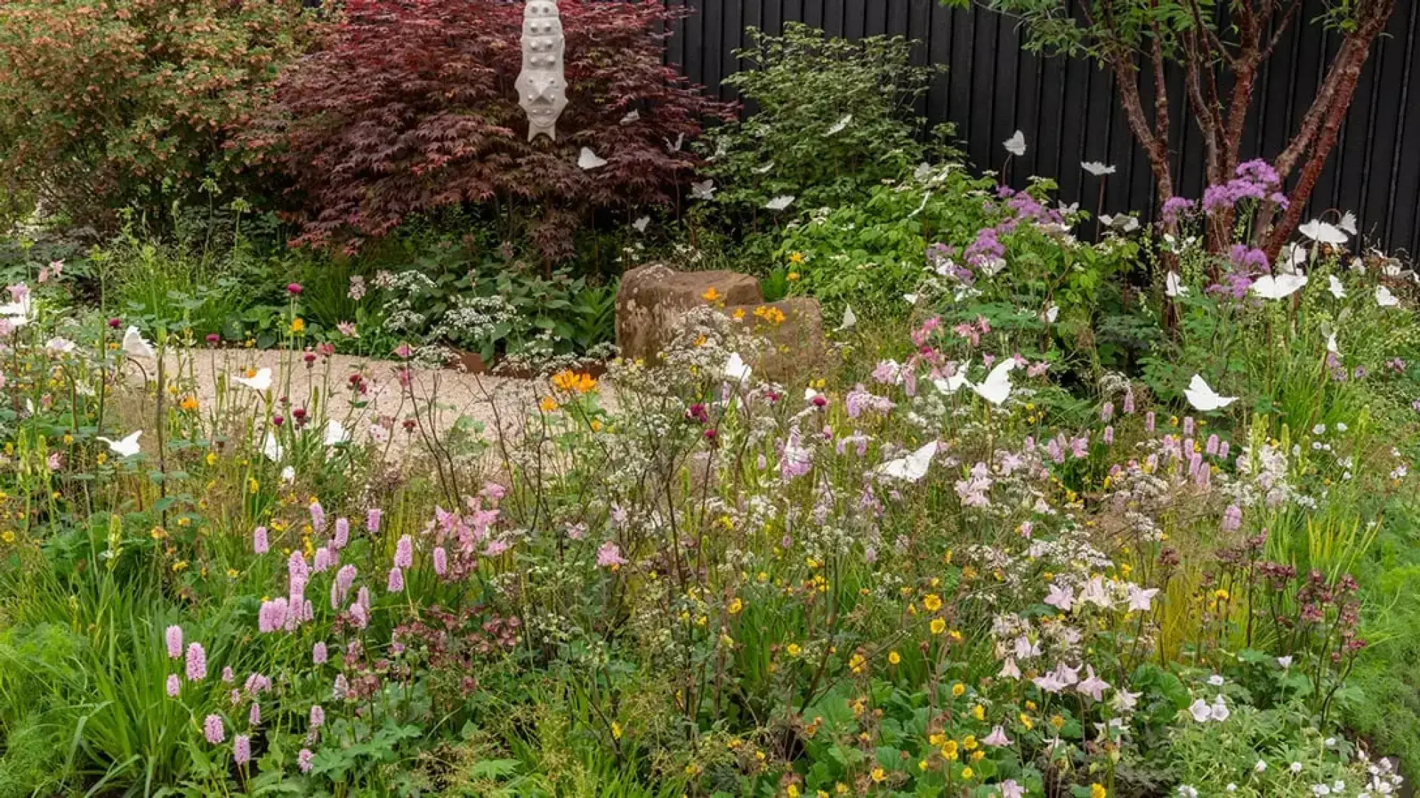 A naturalistic garden filled with pink, yellow, and white flowering perennials, featuring white sculptural butterflies and large natural boulders.