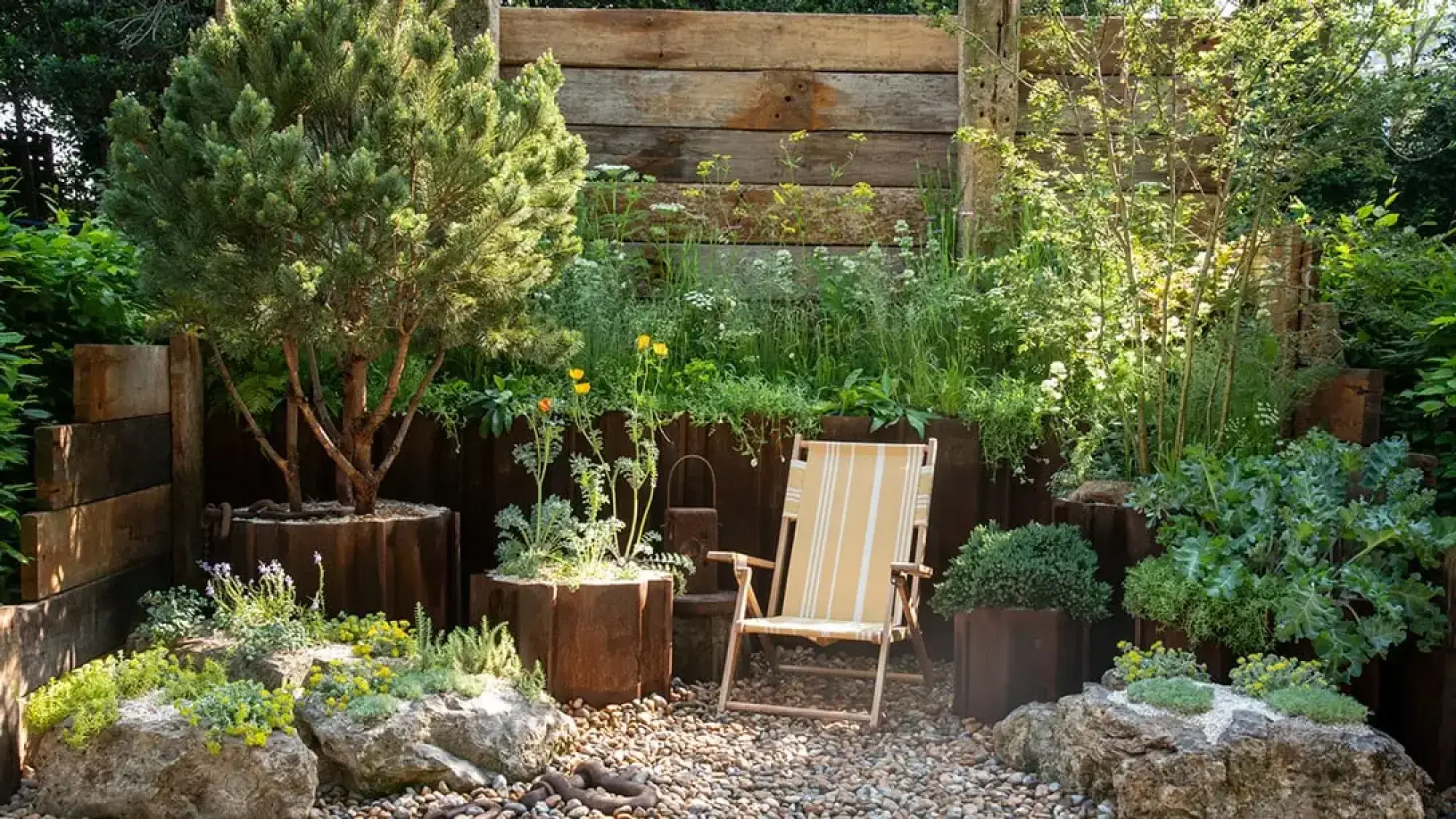 A tranquil container garden with rust-coloured planters, timber wall, gravel, large rocks, and a striped deck chair surrounded by green plants.