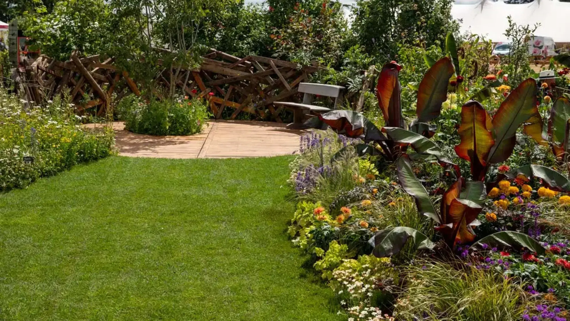 Naturalistic garden with green lawn, wooden deck, bench, rustic log fence, and vibrant red-leafed and mixed flowering plants.