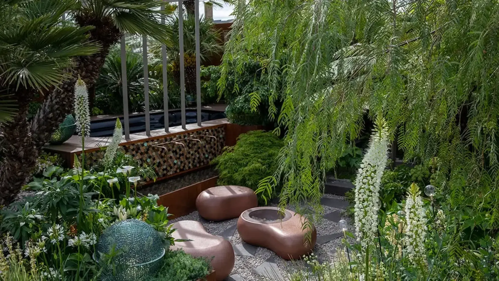 A modern sanctuary garden features organic copper seating amidst lush green foliage, white foxtail lilies, a weeping tree, and a corten steel water feature with a mosaic wall.