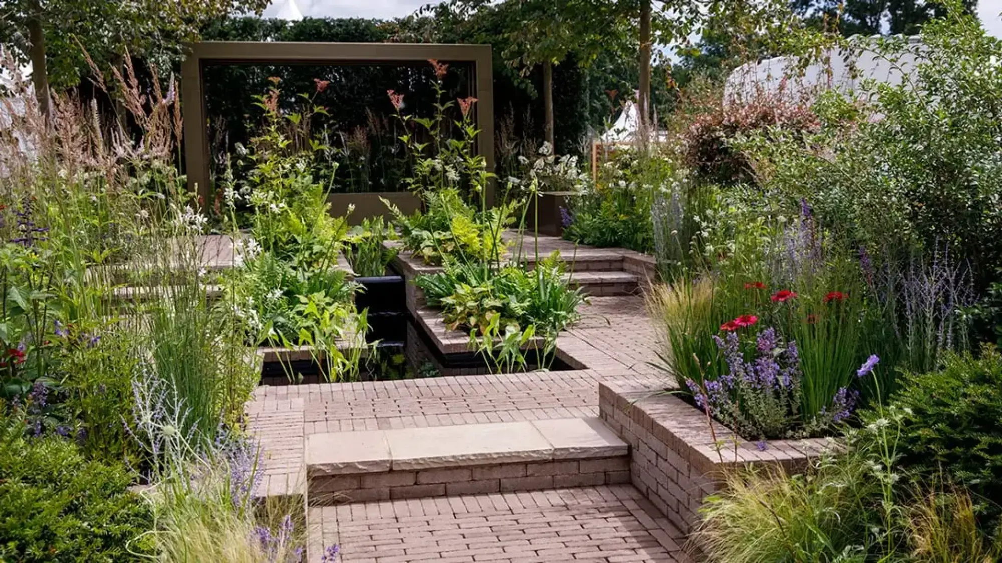 A contemporary show garden features a dark metal arch, tiered water pools, brown brick pathways, and lush planting with ornamental grasses and various colourful perennials.
