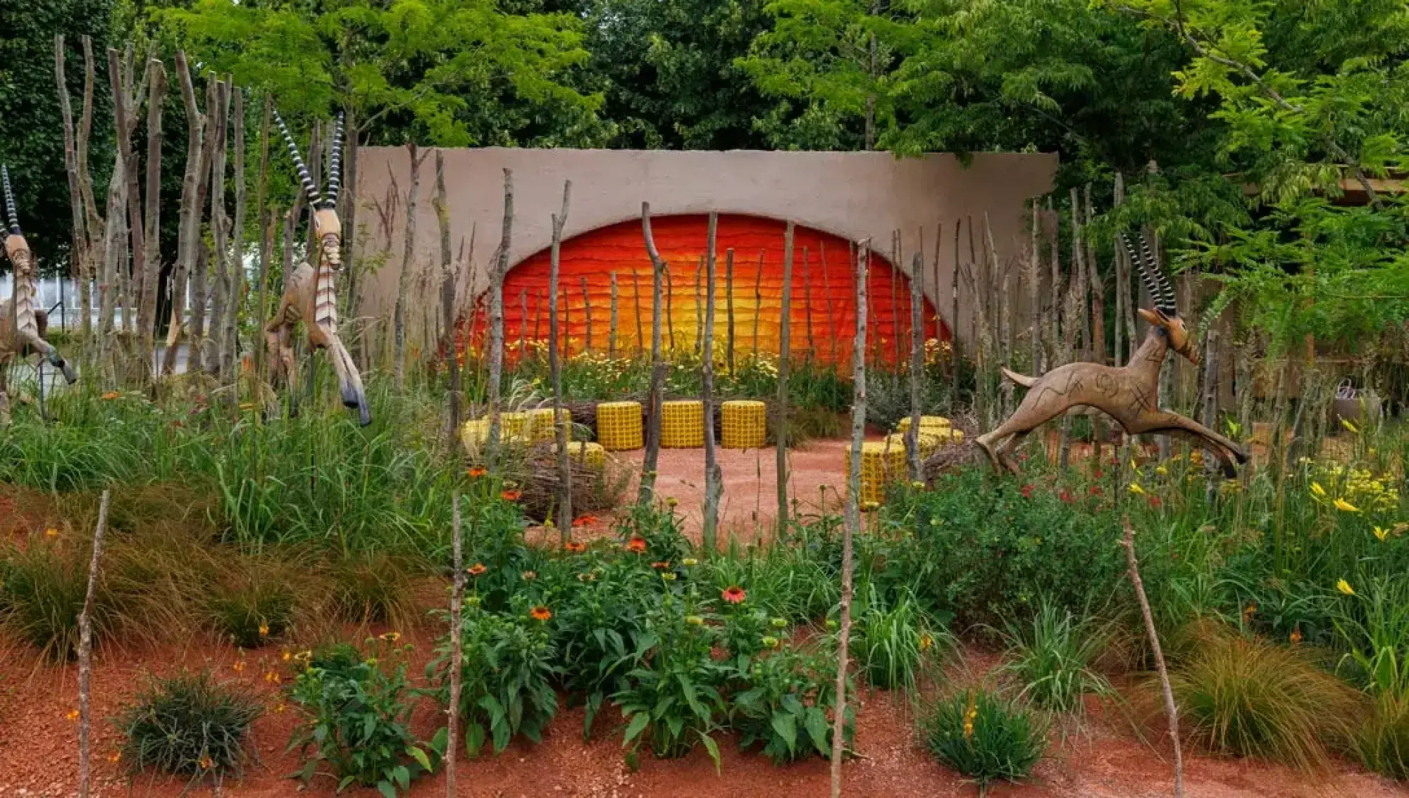 A vibrant savanna garden with a sunrise mural, gazelle sculptures, yellow seating, tall grasses, and colourful flowers on red earth.