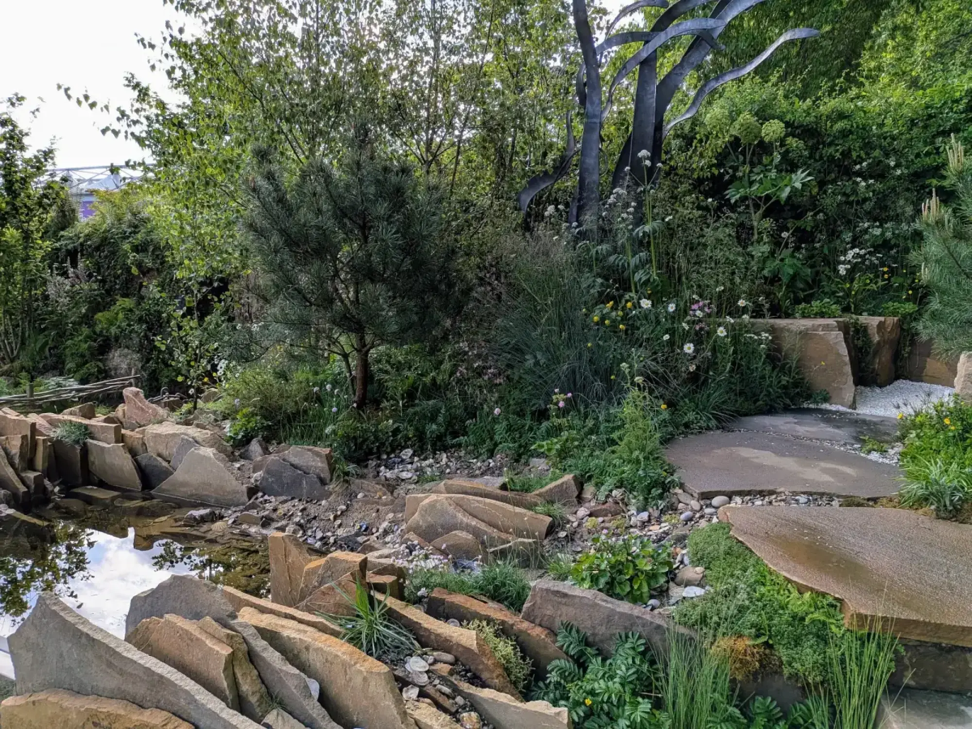 A naturalistic garden with a black metal tree sculpture, irregular stone paving, a shallow water feature, and lush wildflowers and grasses.