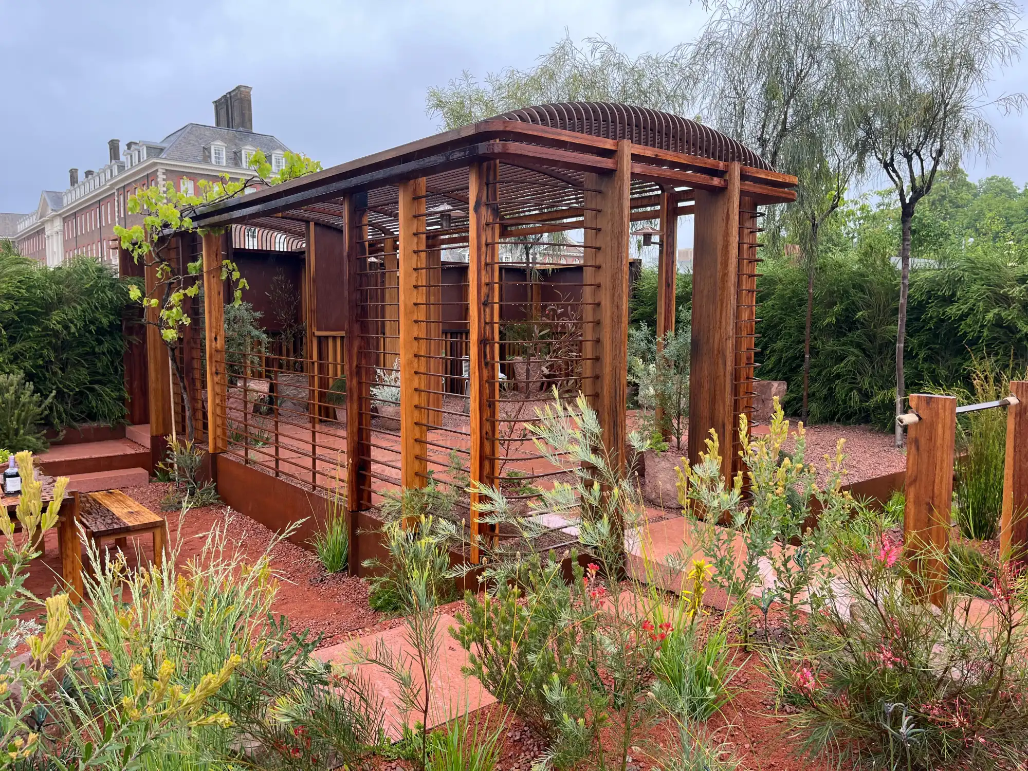 A contemporary Australian arid garden features a large curved timber pergola structure, rust-coloured corten steel walls, red gravel, and native plants.