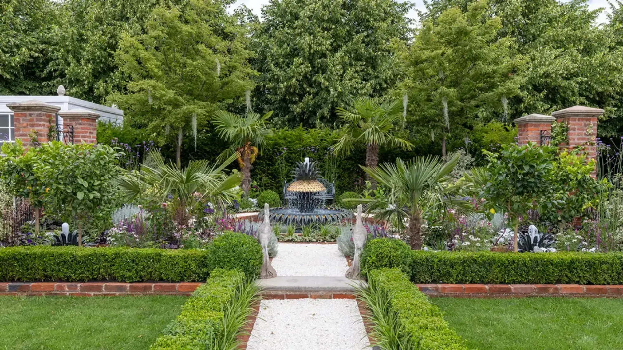 A symmetrical show garden features a prominent pineapple fountain at its center, flanked by crane statues, palm trees, and lush mixed perennial borders, with a white gravel path leading forward.