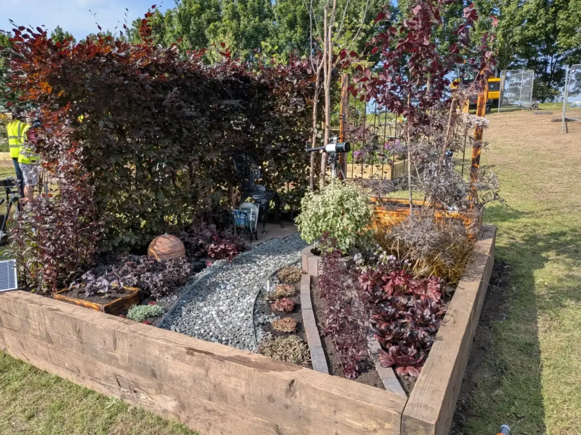 A contemporary show garden with a winding grey gravel path, dark red foliage plants in raised timber beds, a telescope, and a spherical copper ornament.