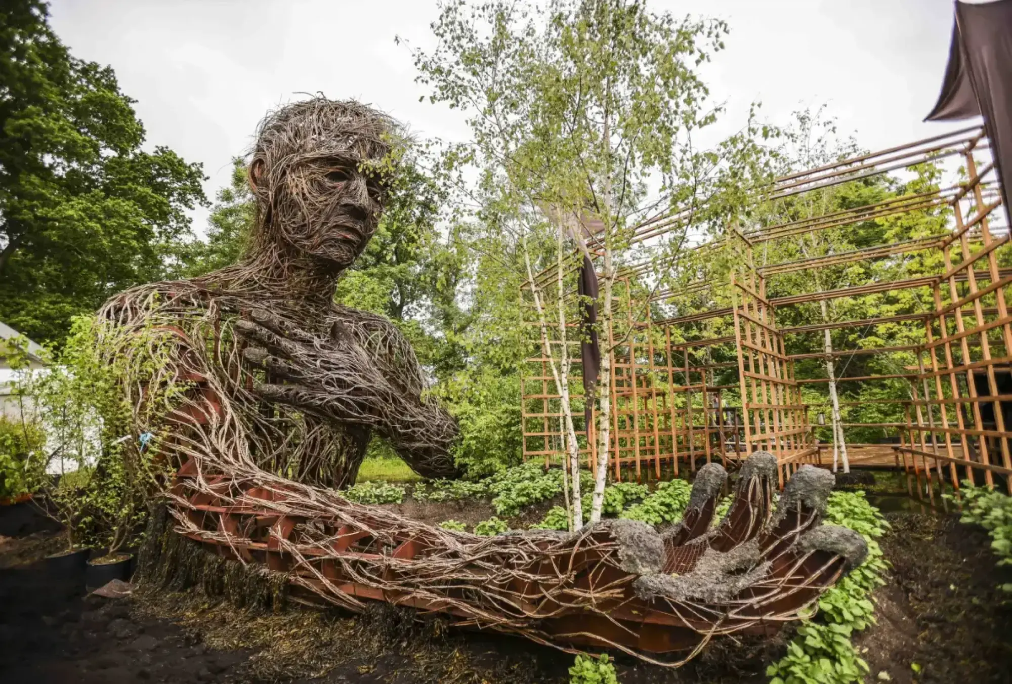A large, seated human figure sculpture, woven from branches and green vines, dominates a naturalistic garden with metal trellises.