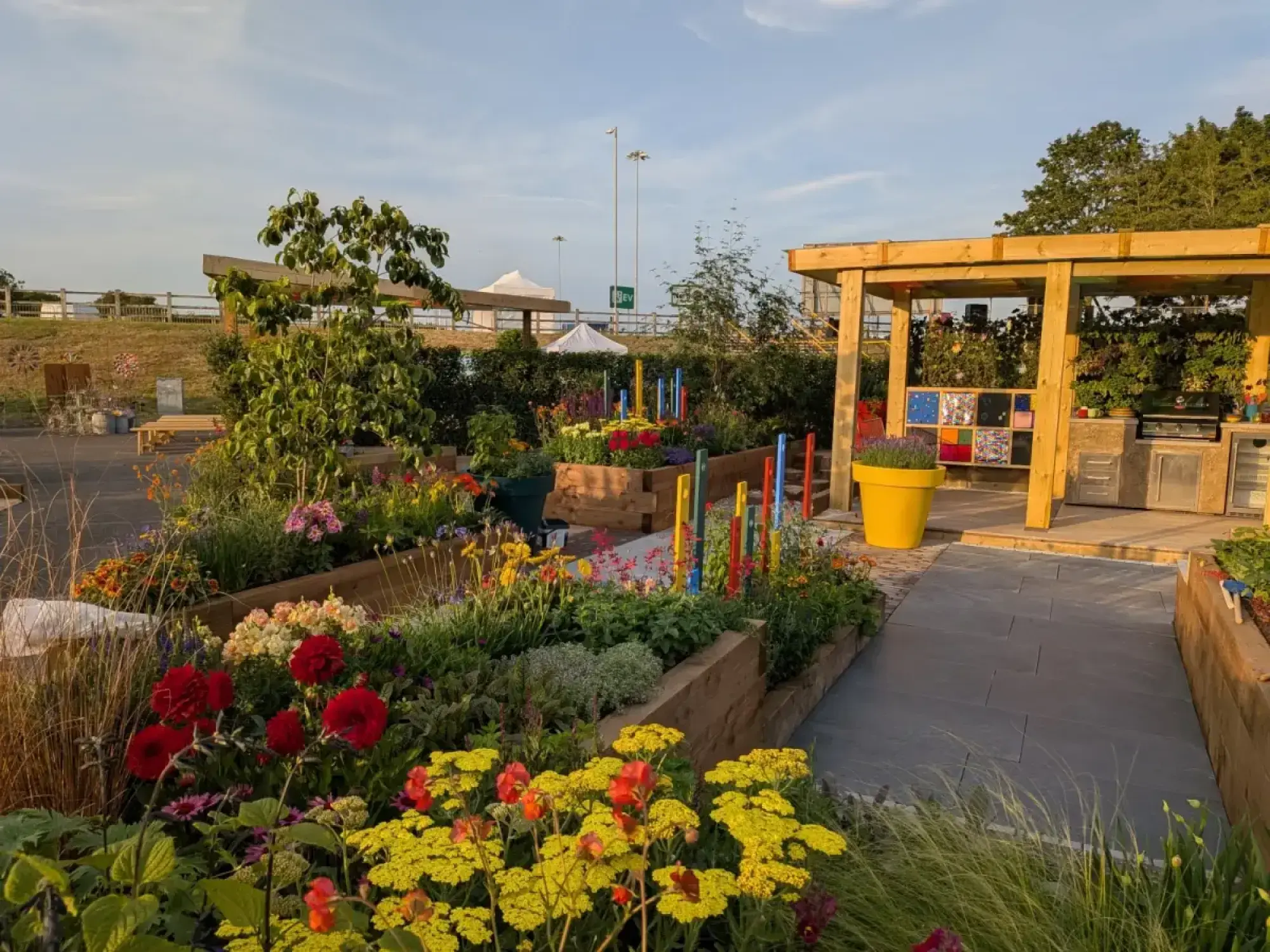 A vibrant show garden features a timber pergola over an outdoor kitchen, colourful mixed perennials in raised beds, and a grey paved path.