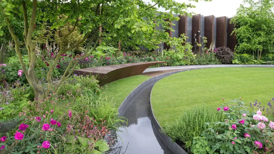 A contemporary show garden featuring a curved rill bordering a manicured lawn, a rust-coloured corten steel bench, and lush planting with pink roses and ferns.