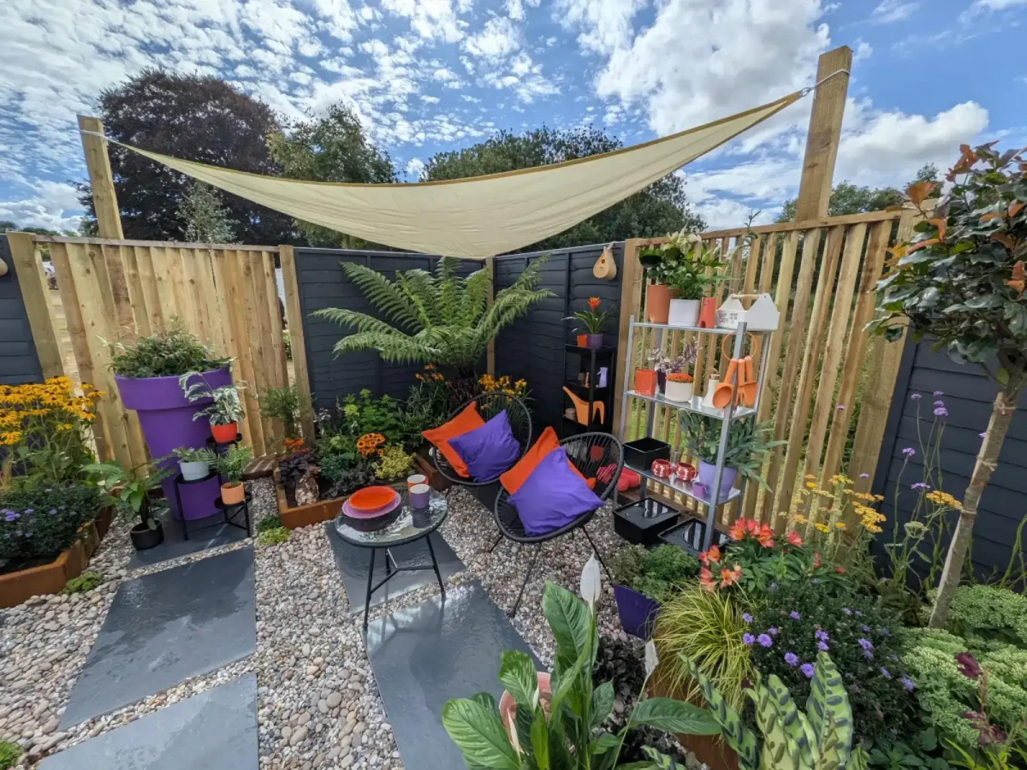 A vibrant small garden with a beige shade sail, black wire chairs with colourful cushions, dark grey paving, gravel, and planting including a large fern.