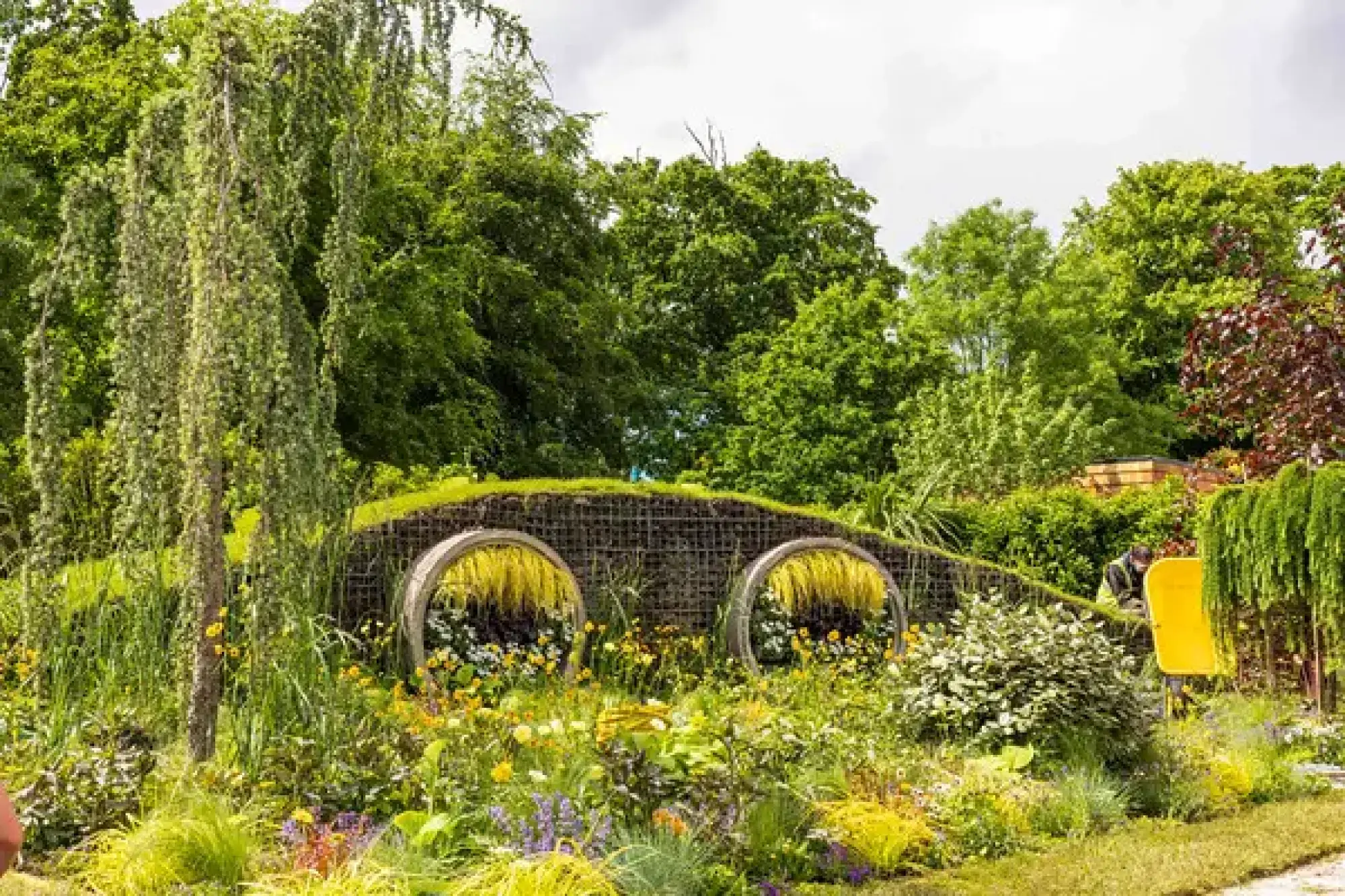 A playful garden features a gabion wall with two round openings, weeping trees, mixed yellow and purple flowers, and a yellow slide.