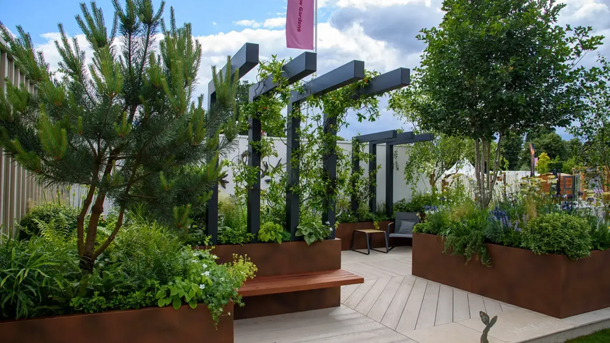 A modern show garden features rust-coloured planters, a dark grey pergola with climbers, an integrated wooden bench, and light grey decking.