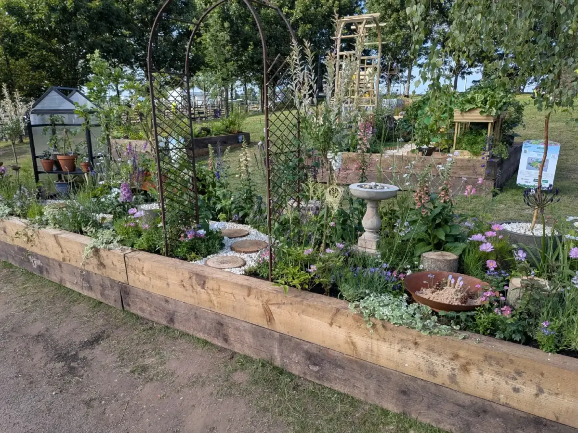 A rustic raised garden border features a mini greenhouse, iron arch, stone bird bath, and wooden beds filled with mixed colourful flowers.