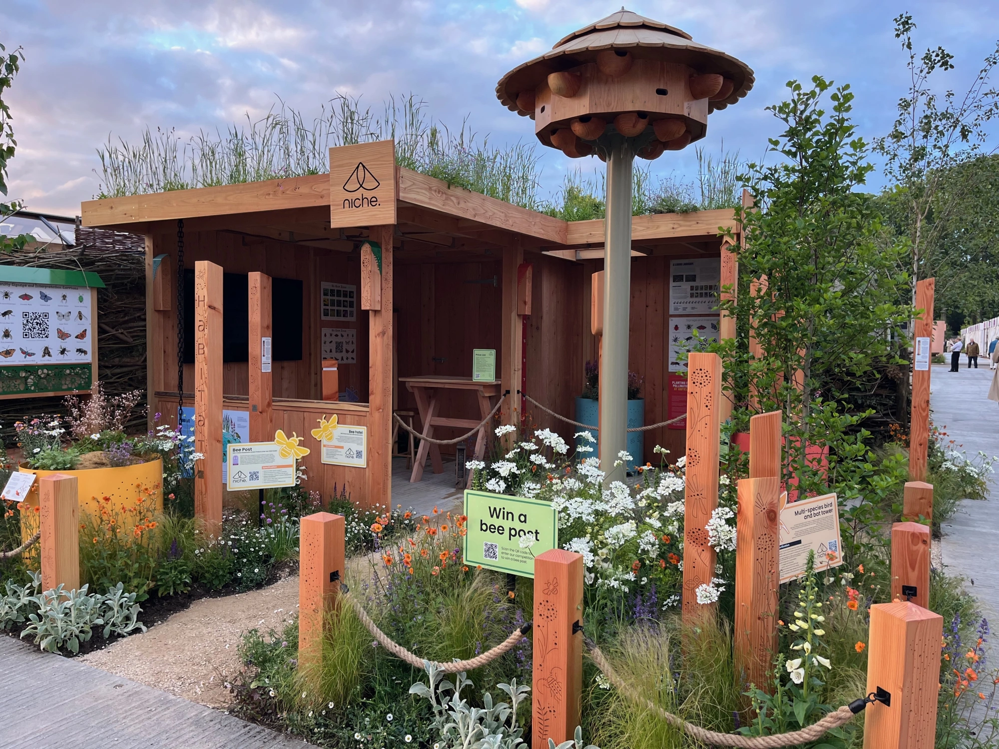 A timber trade stand with a green roof and a multi-tiered wooden bird and bat tower, surrounded by white, orange, and purple wildflowers and carved posts with rope.
