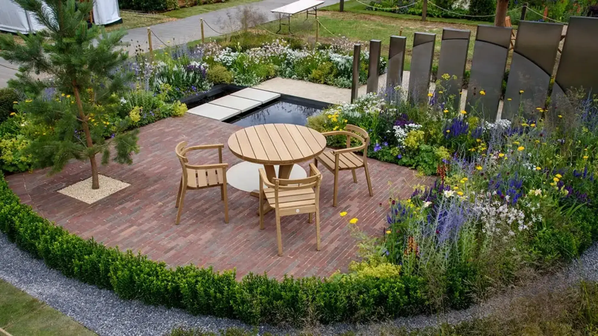 A contemporary show garden features a red brick patio with light wooden dining furniture and a pine tree, surrounded by vibrant mixed herbaceous planting, a pond with stepping stones, and dark curved metal screens.