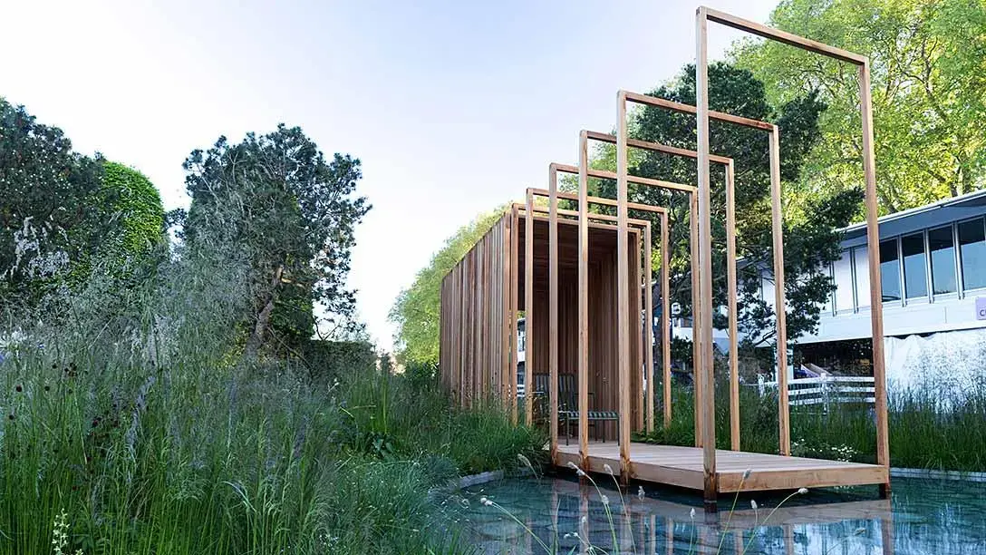 A modern show garden featuring a long timber pavilion with open frames, a wooden walkway over a still water pool, and tall grasses.