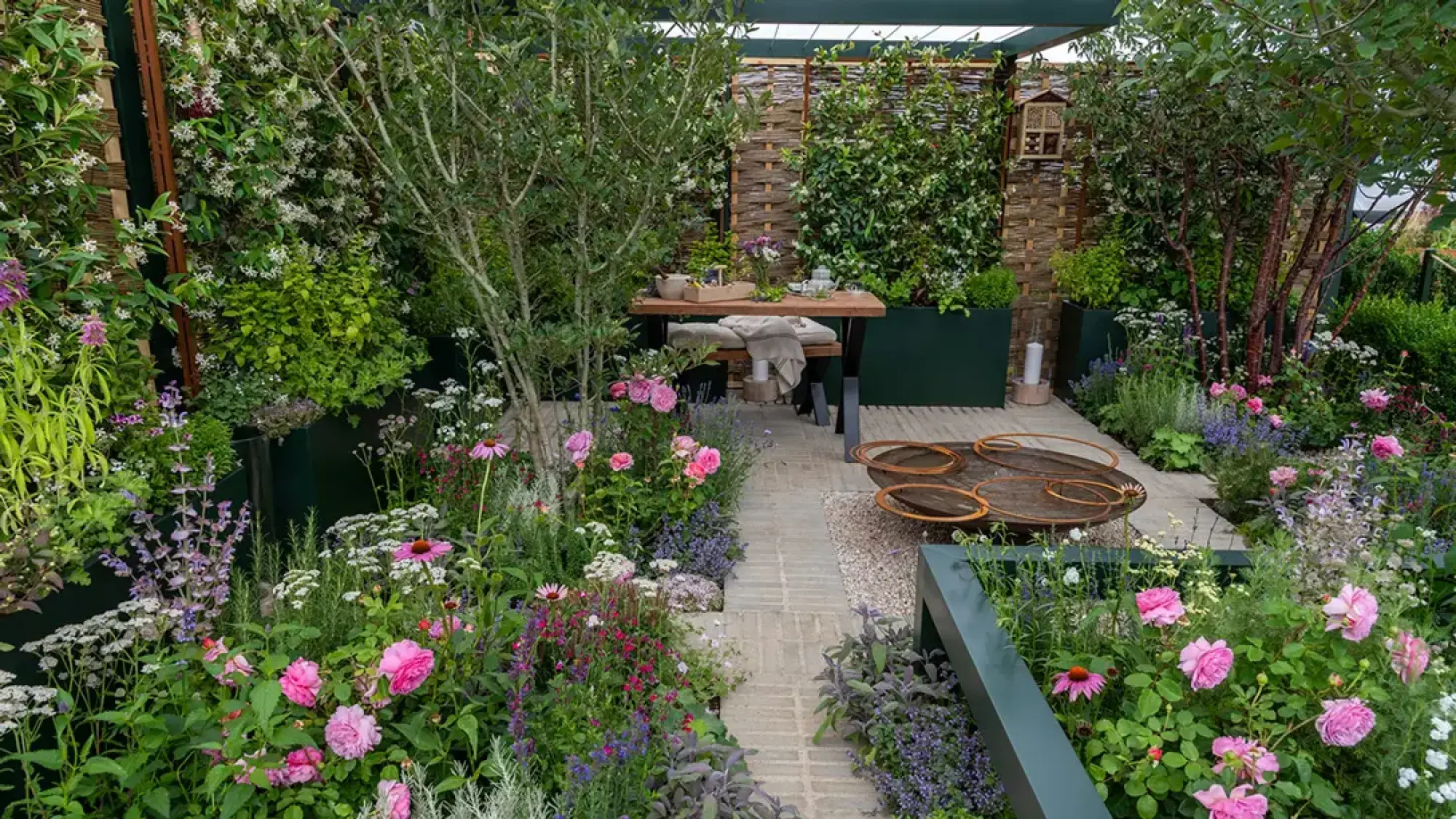 A lush informal garden with a dining table, metal ring sculpture, dark planters, and pink, purple, white flowers.
