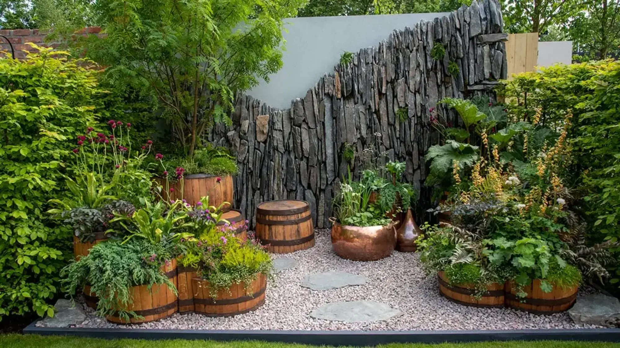 A rustic container garden features a stacked slate wall, wooden barrel planters, copper pots, and lush green and flowering plants.