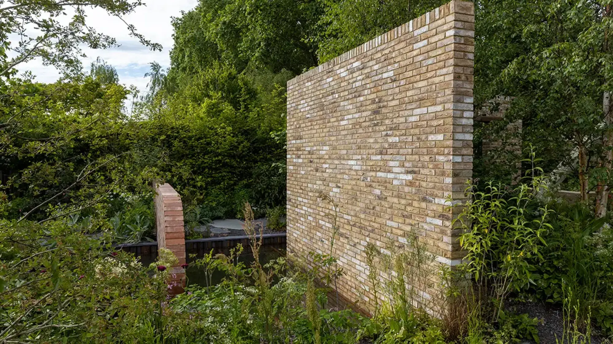 A contemporary show garden featuring a tall pale brick wall, a curved terracotta brick wall, a dark water pond, and lush green naturalistic planting with slender trees.
