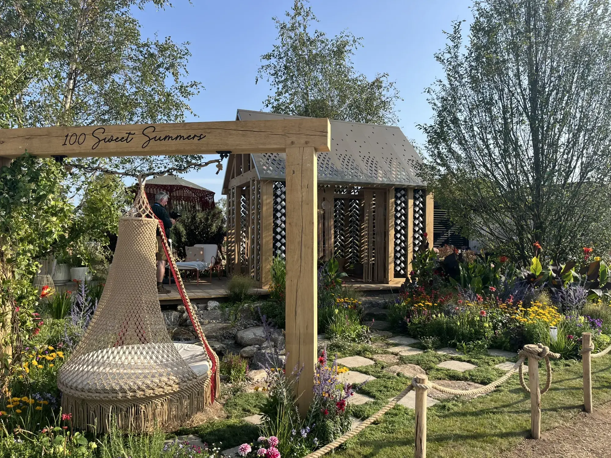 The '100 Sweet Summers' garden features a wooden arch, a macrame daybed, and a patterned timber pavilion surrounded by colourful flowers.