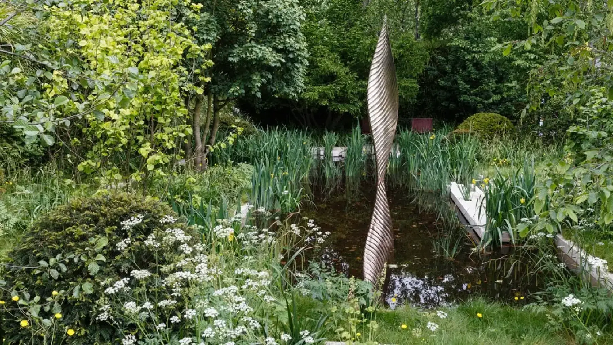 A tranquil water garden showcases a tall, spiraling bronze sculpture amidst lush green foliage, aquatic plants, and clusters of white and yellow wildflowers.