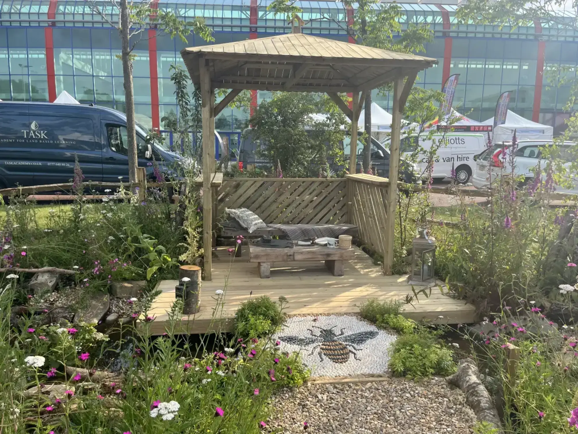 A naturalistic showcase garden features a wooden gazebo, a stream, and a wildflower meadow with a prominent bee pebble mosaic on the timber deck.