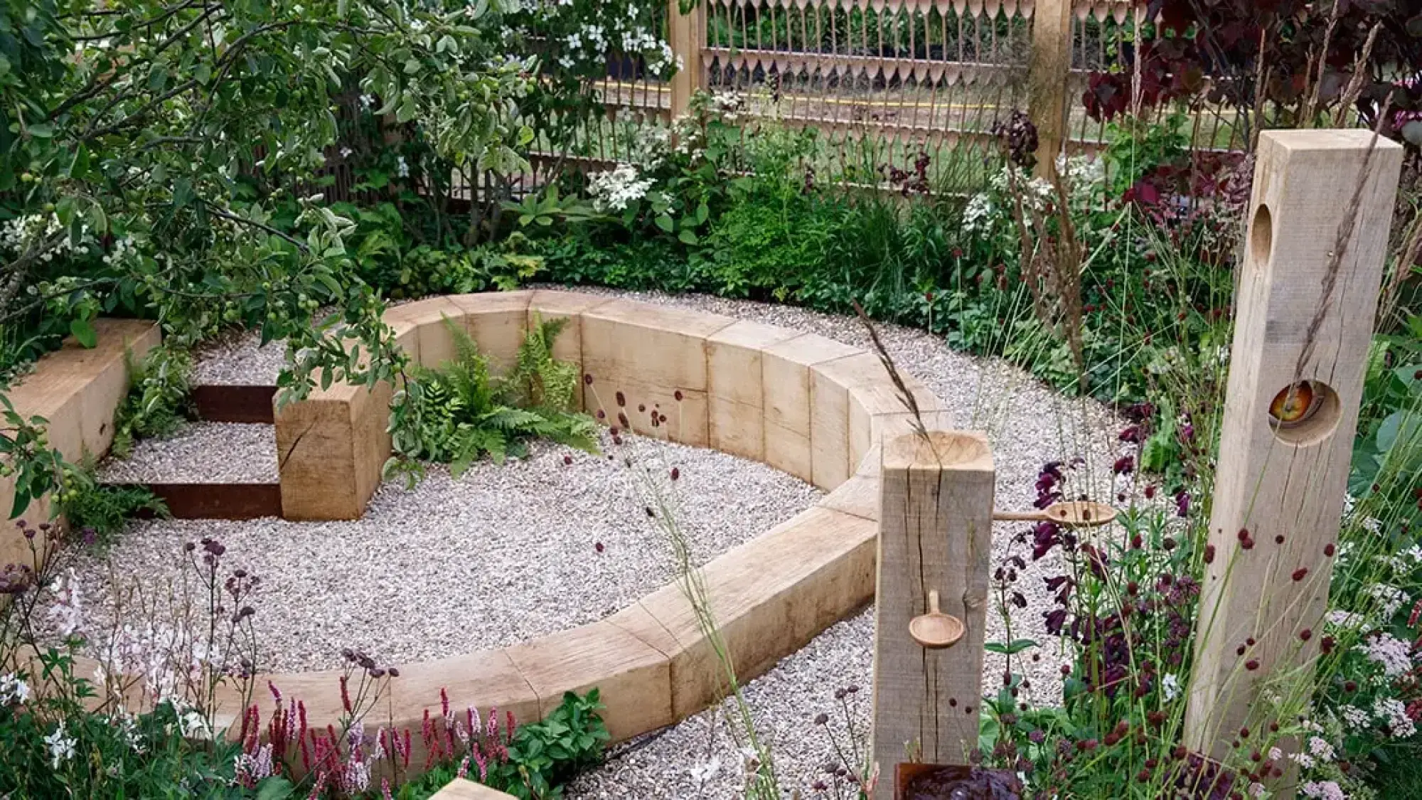 A naturalistic Get Started Garden features a curved timber bench set in gravel, with carved wooden posts holding spoons and flowering plants.