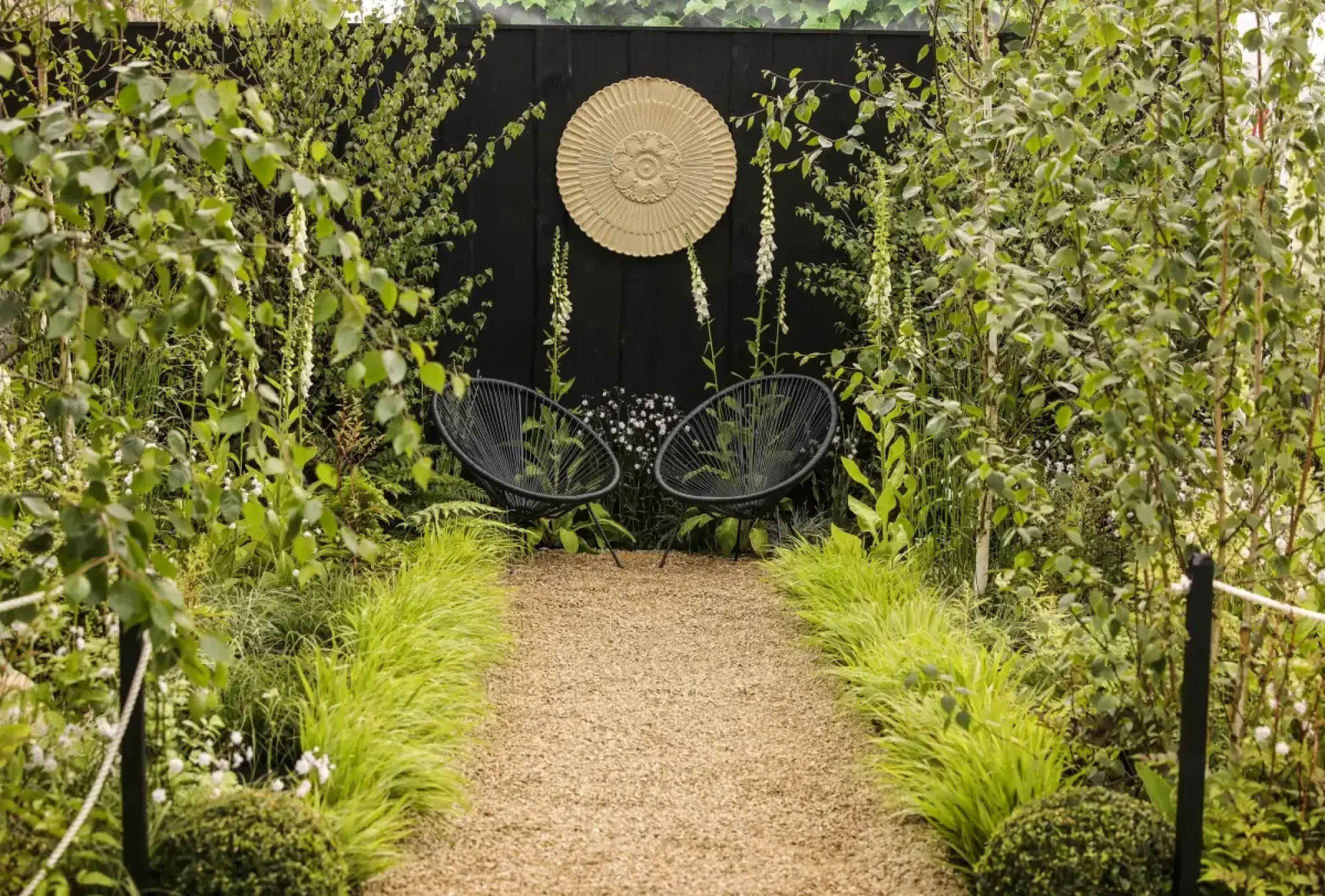A gravel path leads to black wire chairs against a black wall with a gold sculpture, surrounded by green trees and white foxgloves.