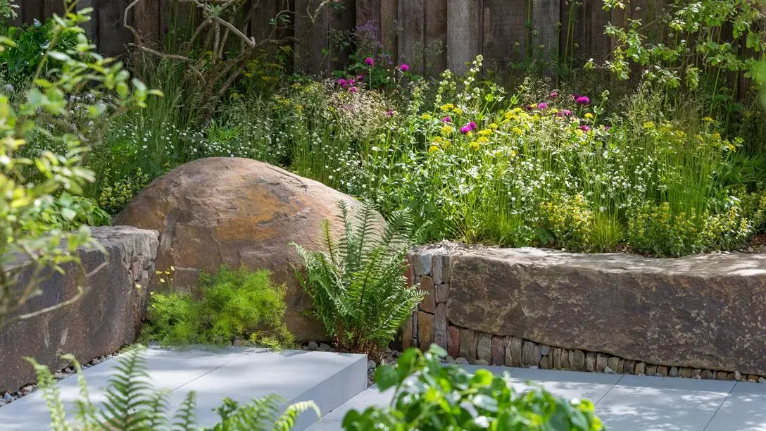 A naturalistic garden features large weathered boulders, dry-stacked stone walls, light grey paving, and lush ferns, grasses, and colourful meadow flowers.