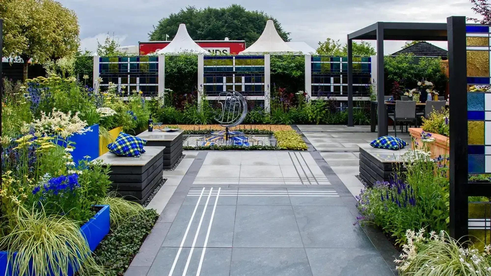 A modern show garden features a '5D' sculpture, geometric grey paving, colourful planters, vibrant perennials, and a black pergola with outdoor dining.