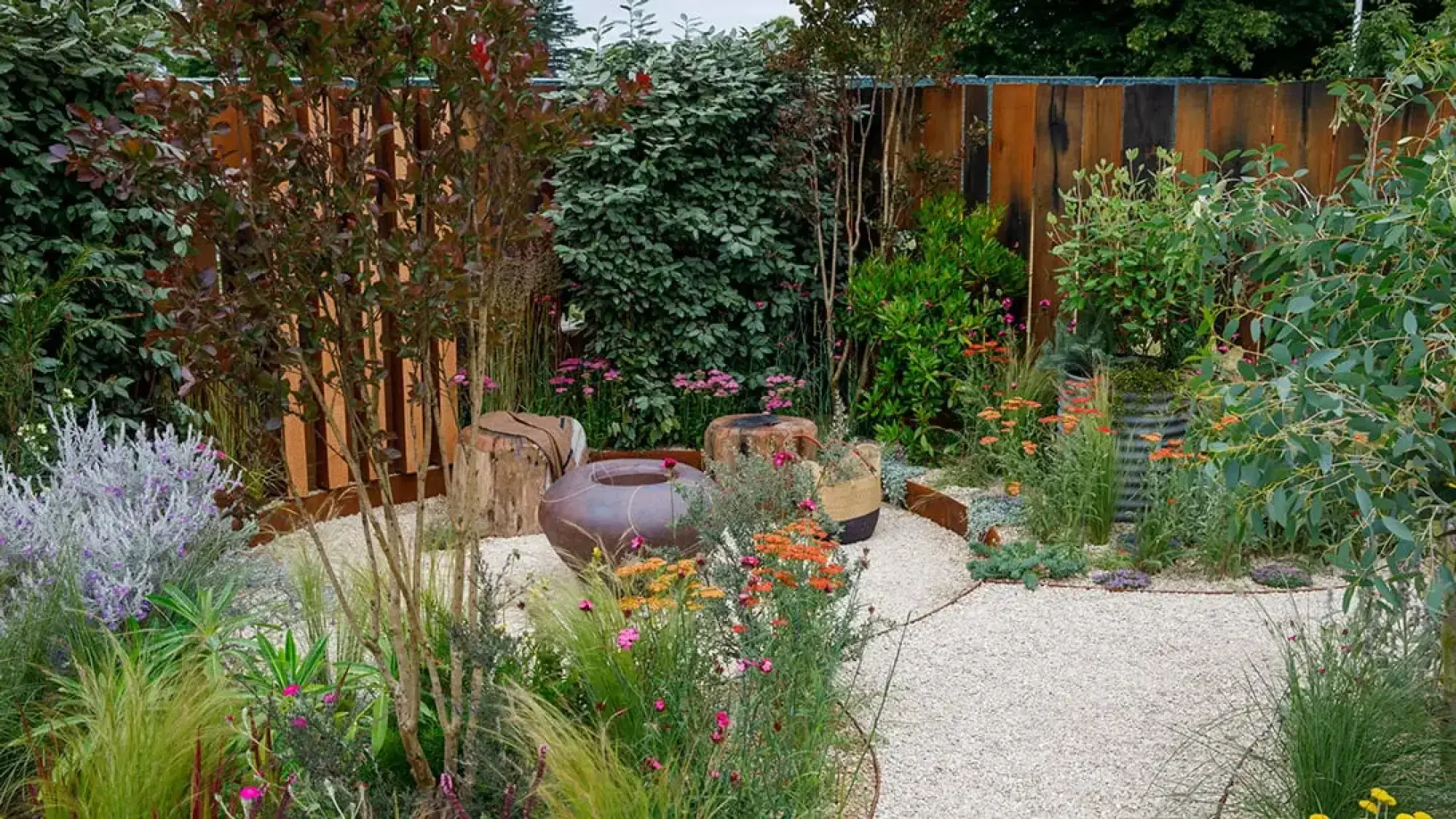 A naturalistic garden with a large brown pot, timber log seats, and a light gravel path, featuring orange and pink flowers, ornamental grasses, and silvery shrubs, against a weathered timber fence.