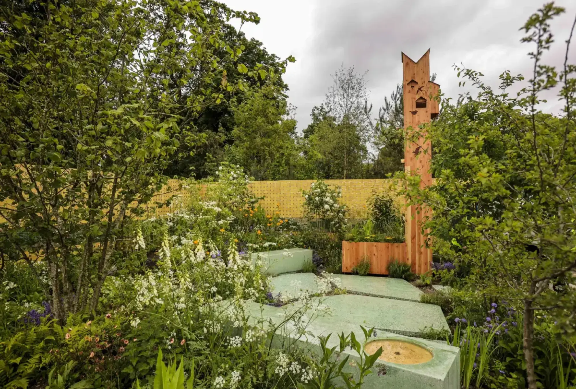 A modern ecological concept garden features a tall wooden habitat tower, light green recycled concrete paving with a water basin, and naturalistic planting against a yellow woven timber screen.