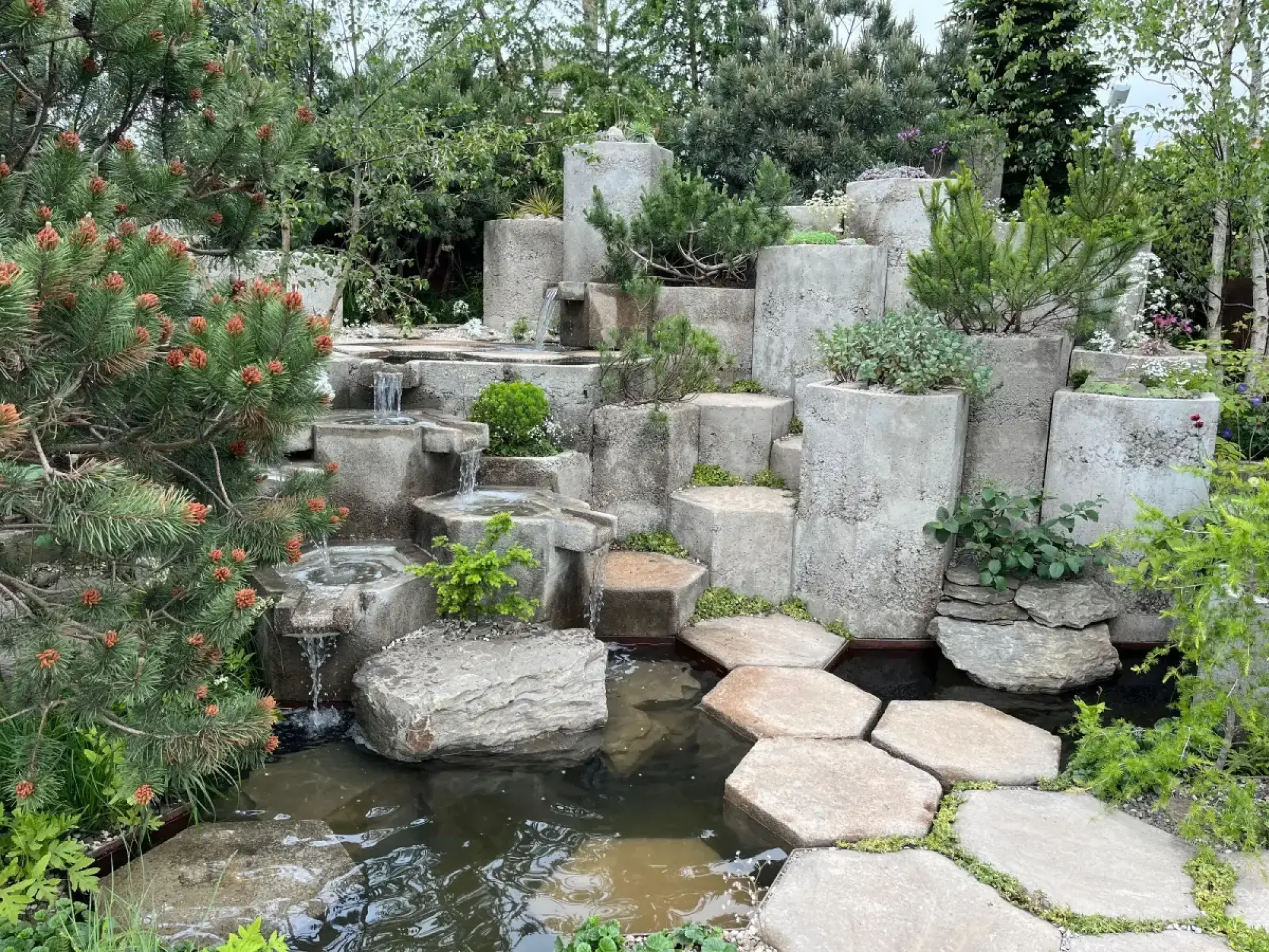A tranquil small show garden features a tiered concrete waterfall and pond with hexagonal stepping stones, surrounded by dwarf pines and green foliage.