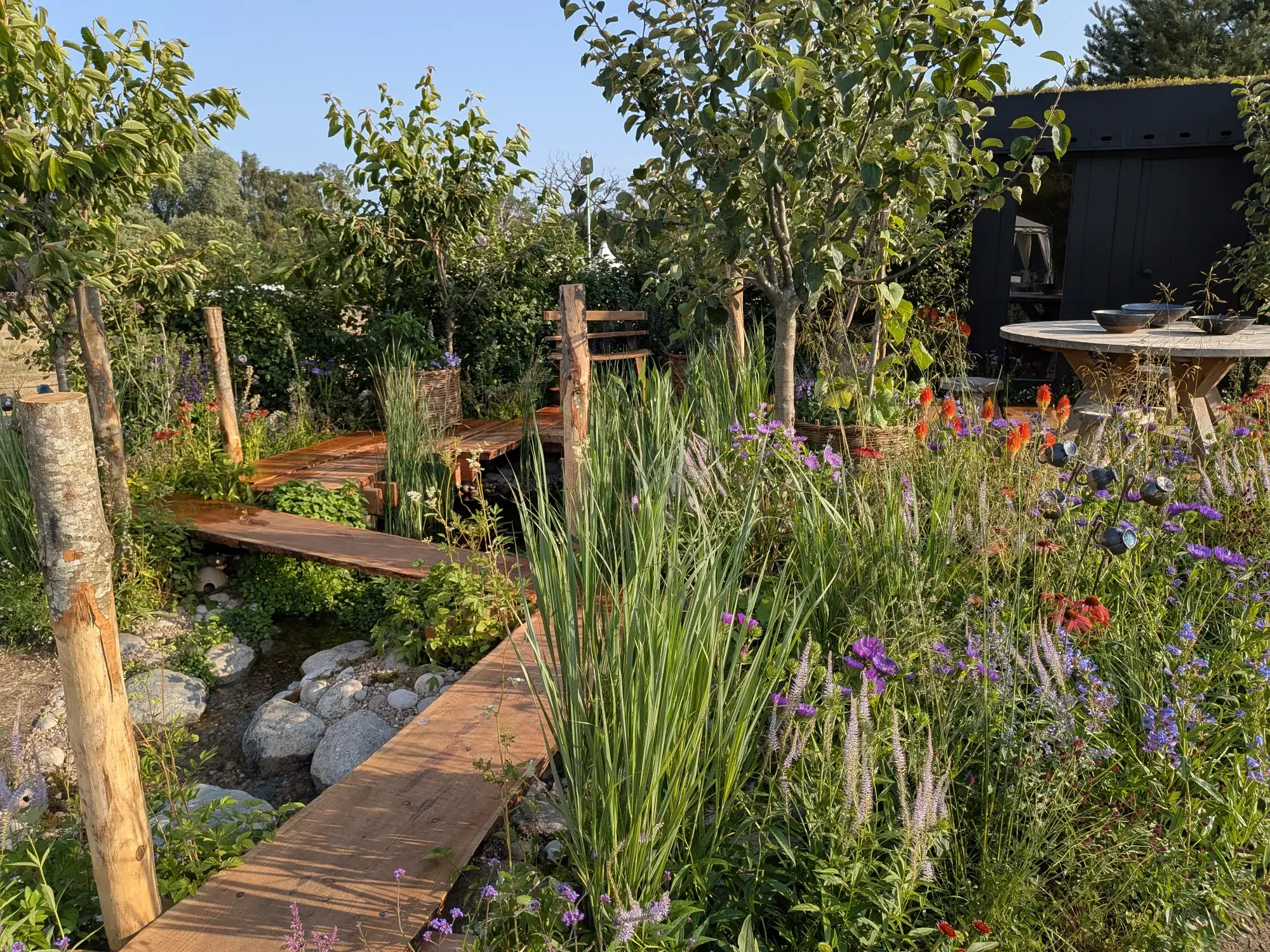 A naturalistic show garden with a winding wooden path over a rocky stream, fruit trees, mixed perennial planting, a round wooden dining table, and a black timber cabin with a green roof.