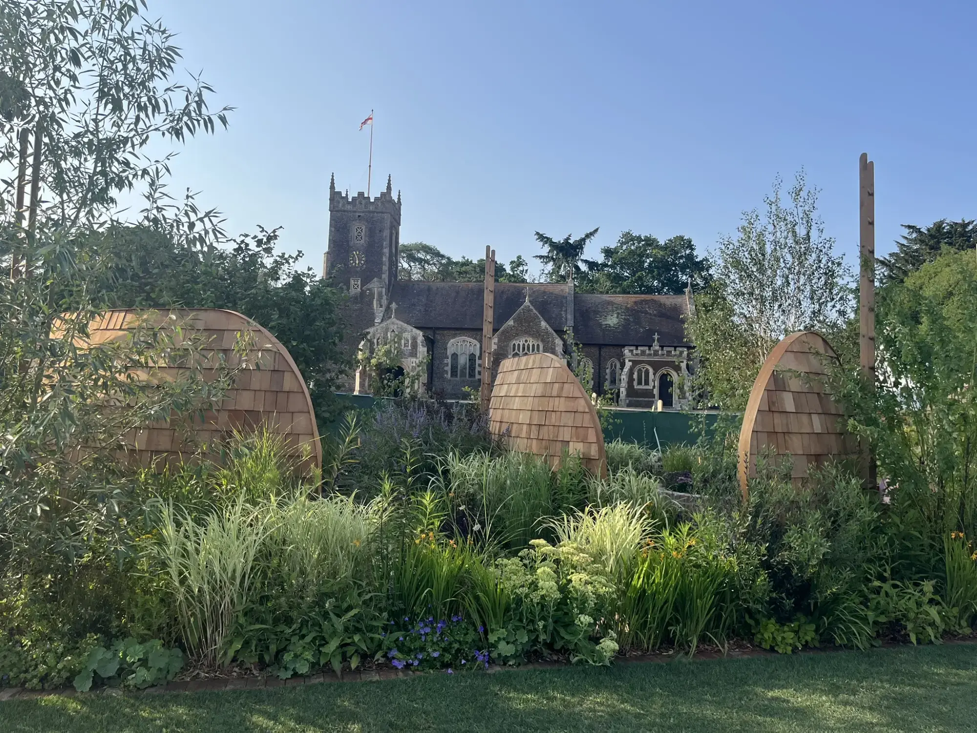 A naturalistic show garden features three large, curved wooden pods surrounded by lush green grasses, mixed perennials, and purple and yellow flowers.