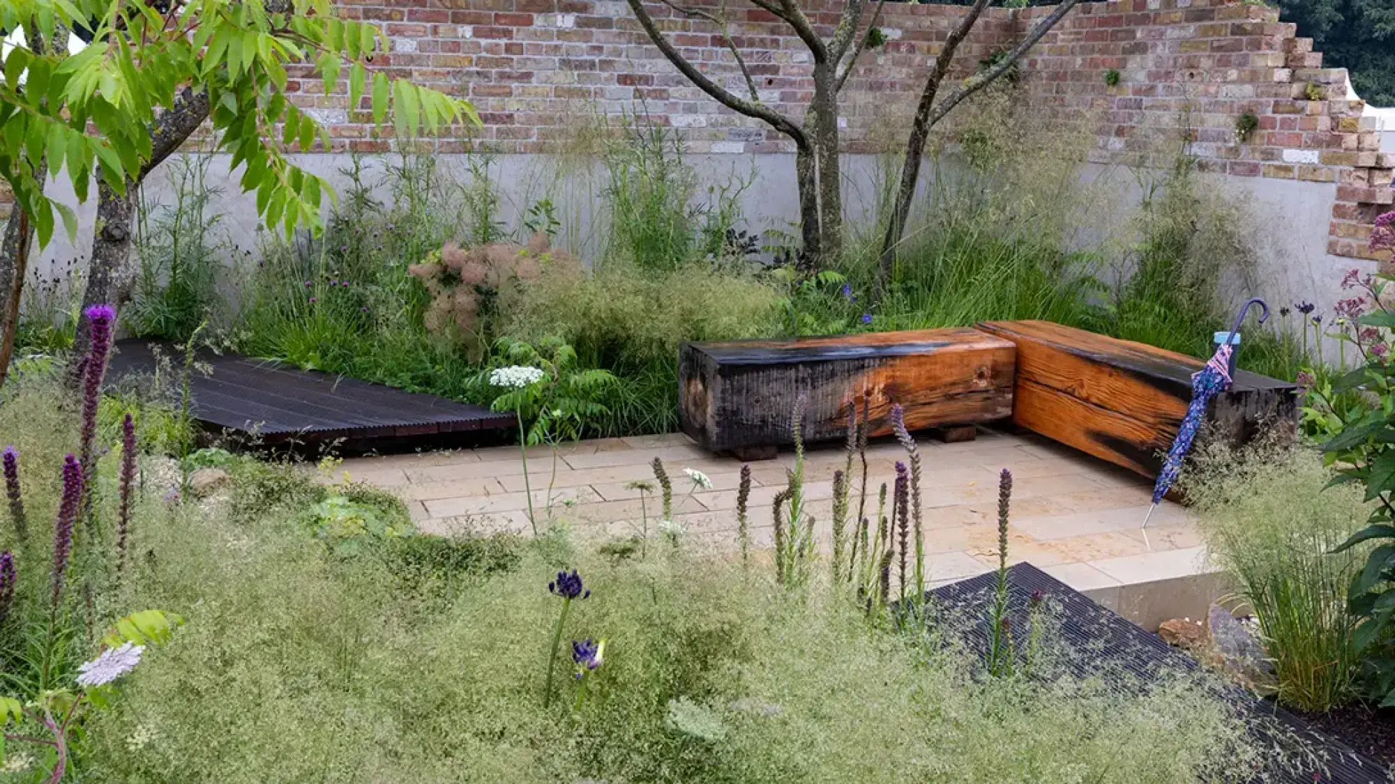 A contemporary naturalistic garden features charred timber benches, a corrugated platform, pale paving, feathery grasses, and purple and white flowers.