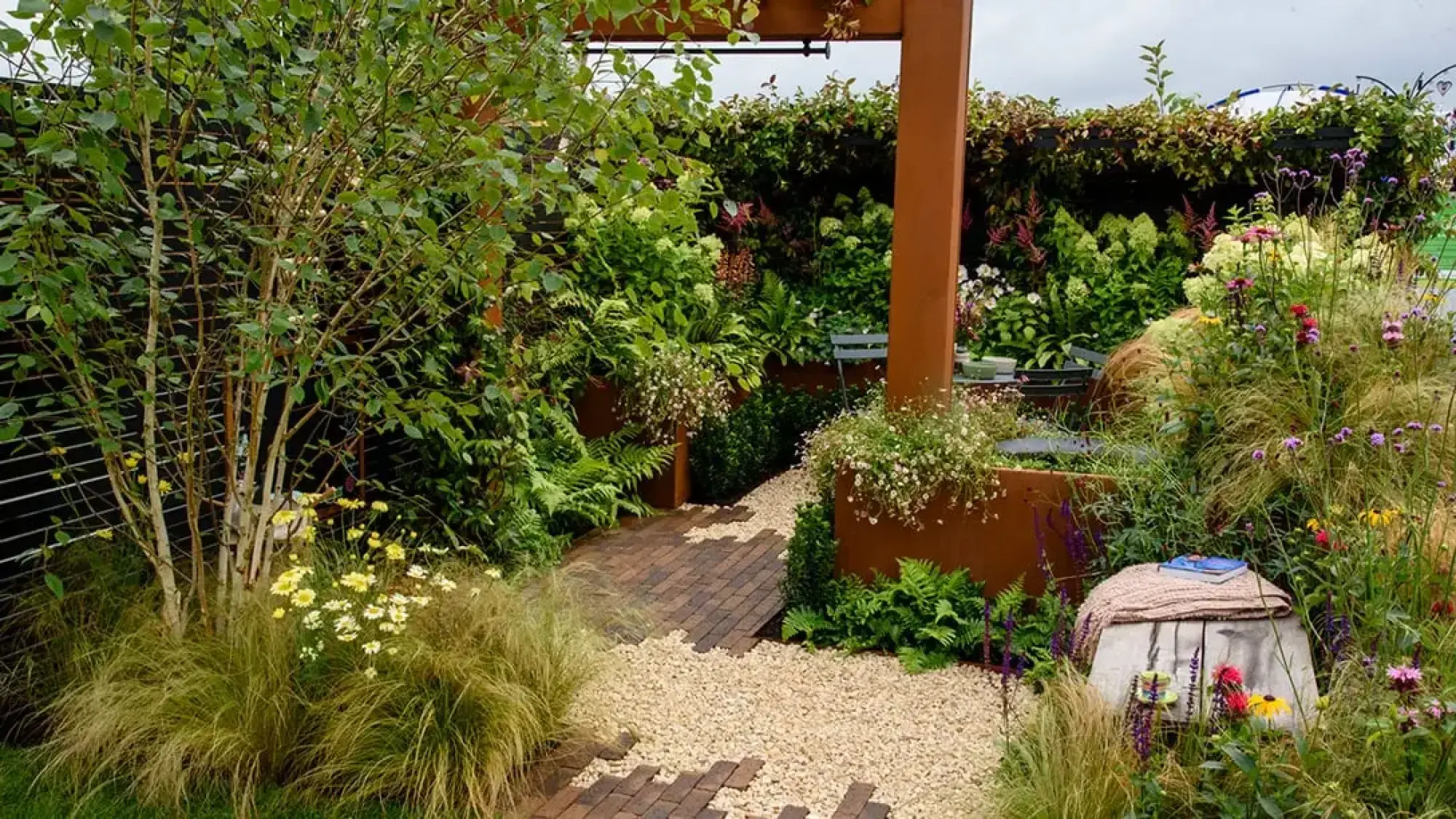 A lush terrace garden features prominent Corten steel planters and a pergola, a winding path of brick and gravel, and abundant naturalistic planting including a multi-stemmed birch tree.
