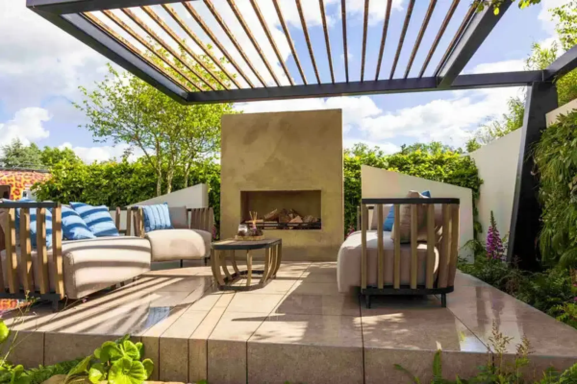 A modern outdoor lounge garden with a slatted pergola, rendered fireplace, beige paving, sofas, and green plants.