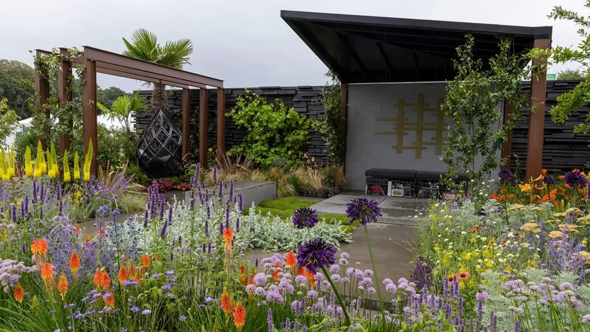 A contemporary show garden features a black geometric hanging chair under a Cor-Ten steel pergola and a modern black shelter, surrounded by vibrant purple, orange, and yellow prairie-style planting.