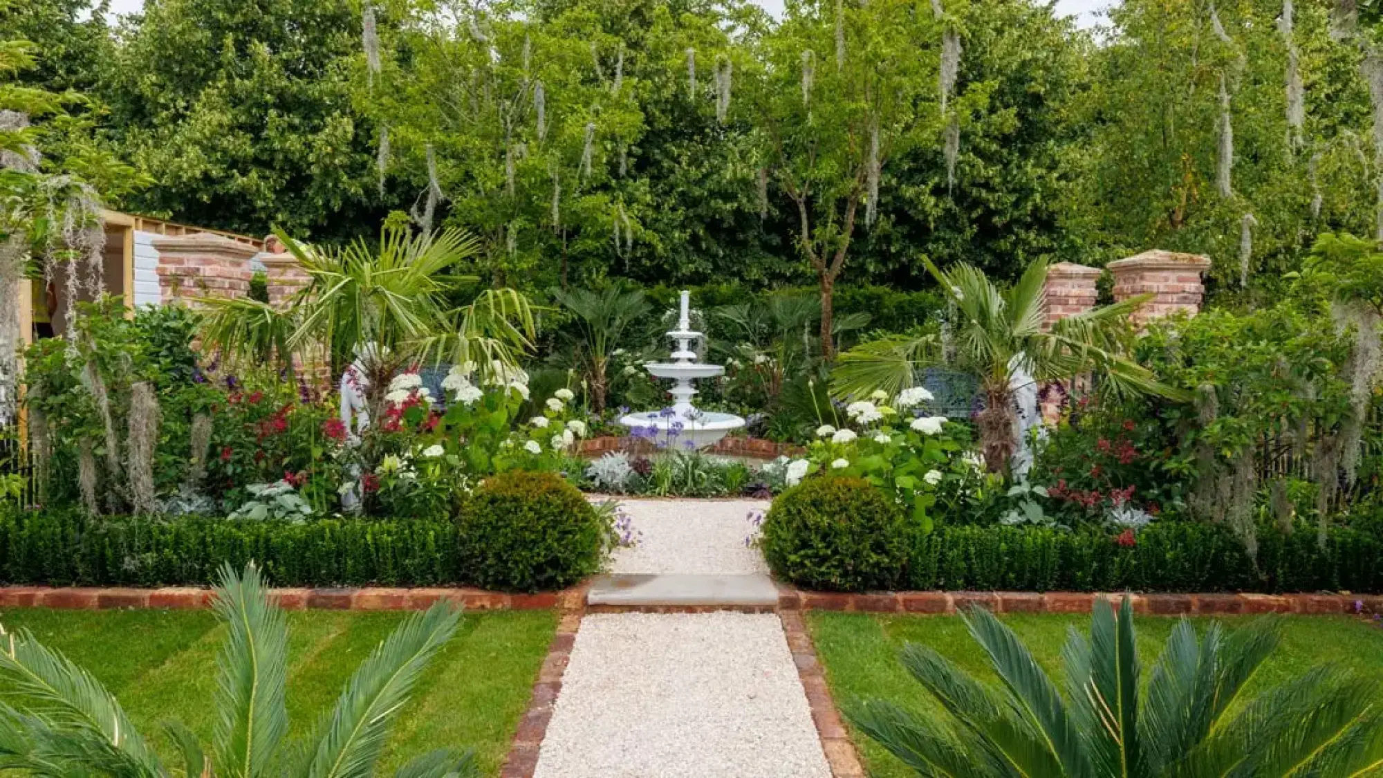 A formal garden with a white tiered fountain, lush palm trees, white hydrangeas, Spanish moss, and a central gravel path.