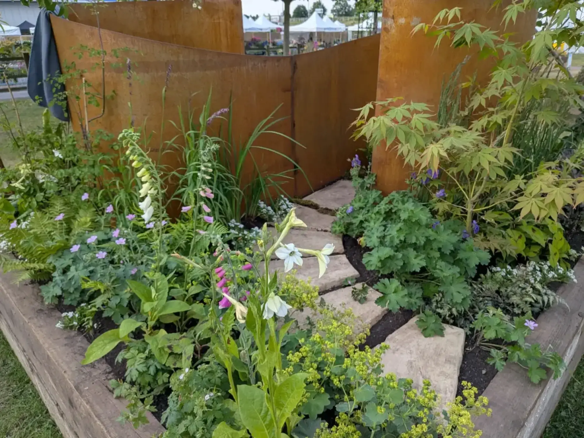 A lush, tranquil garden border with rust-orange Corten steel screens, a flagstone path, and green plants featuring white, pink, and purple flowers.