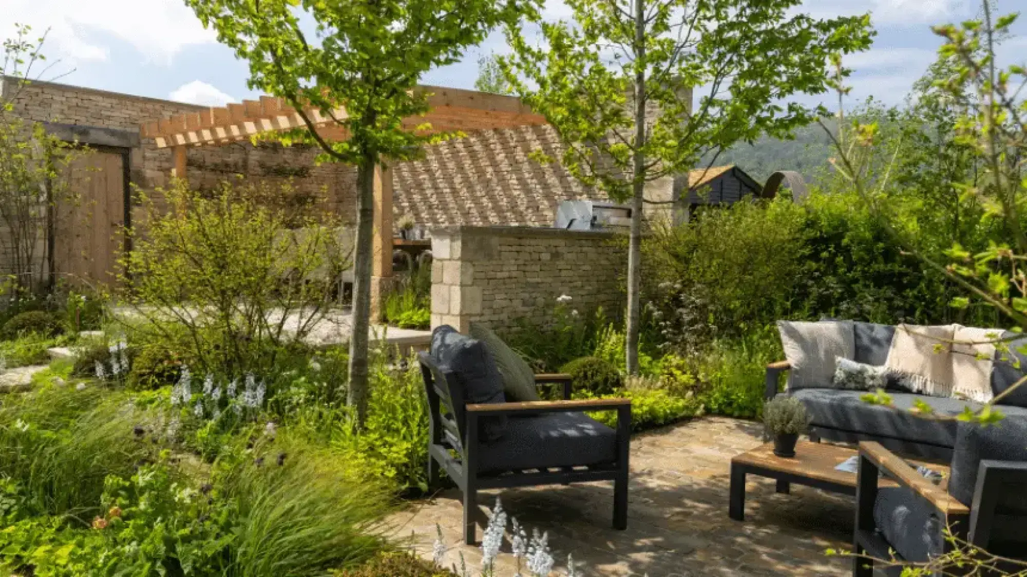 A contemporary Cotswold garden features a stone wall, wooden pergola, and outdoor kitchen, with modern lounge seating on a paved patio surrounded by lush green planting.