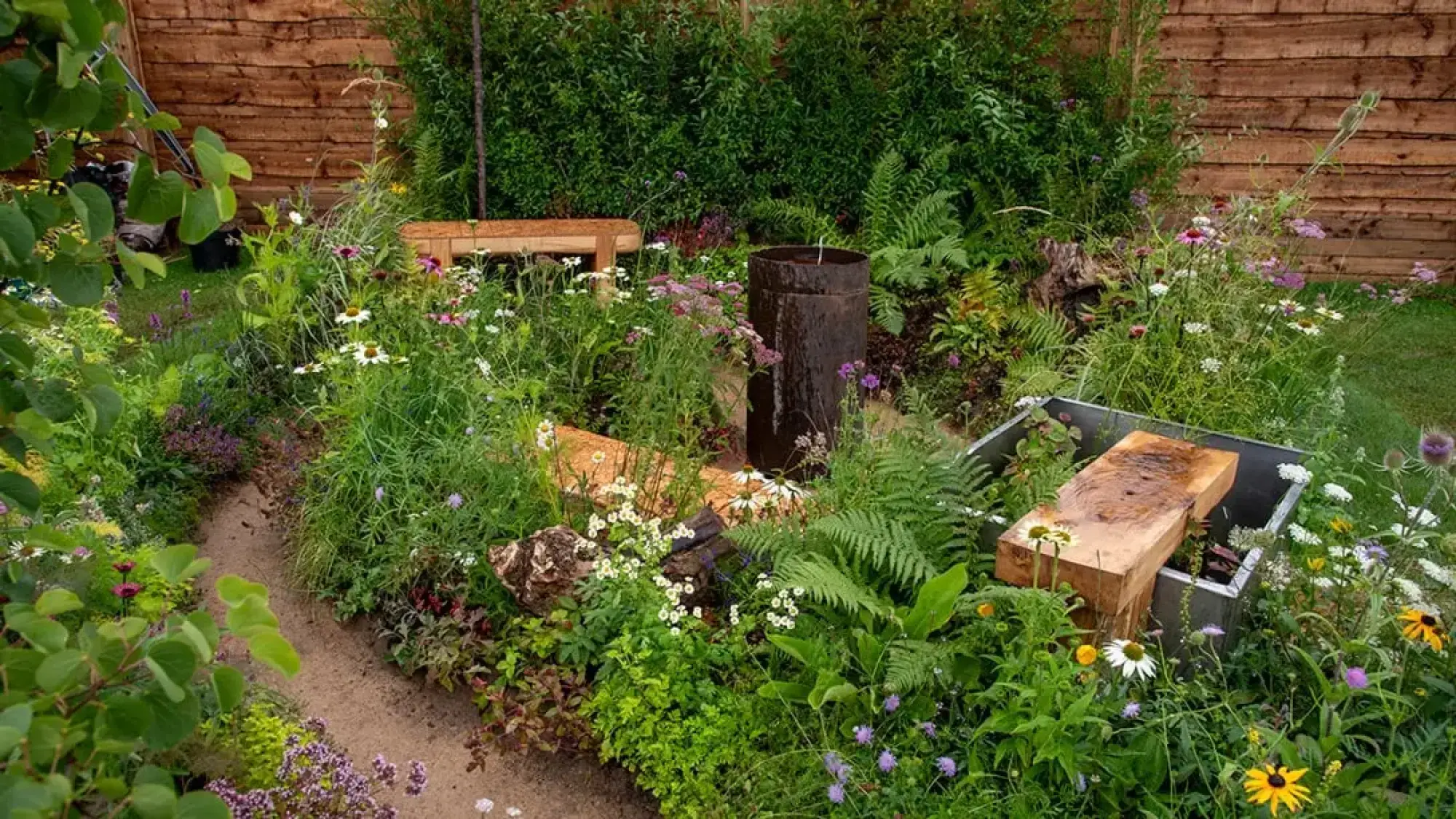 A naturalistic terrace garden with a dark cylindrical water feature, chunky timber seating, and a sandy path winding through abundant wildflowers and ferns.